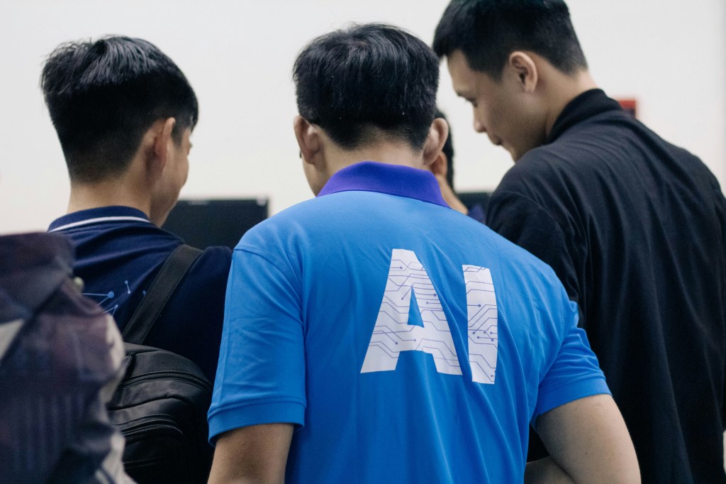 Gruppe von drei Menschen. Einer in der Mitte hat AI am Rücken seines T-Shirts stehen.
